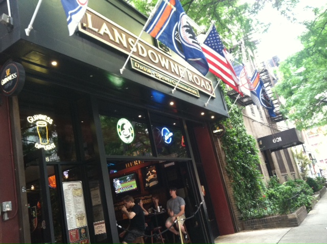 Exterior of Lansdowne Road Irish pub in Hell’s Kitchen, NYC, with flags out front, open windows, and locals inside watching a game — classic New York neighborhood bar scene.